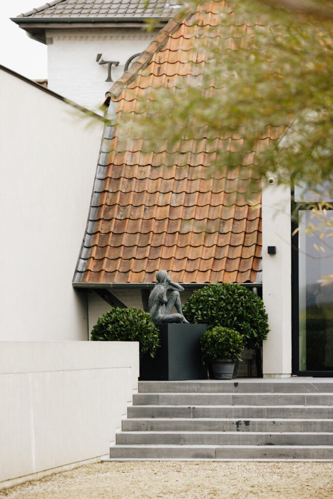 Buitensculptuur en groene plantvakken aan de trap bij de ingang van ’t Convent, met zicht op het karaktervolle dak.